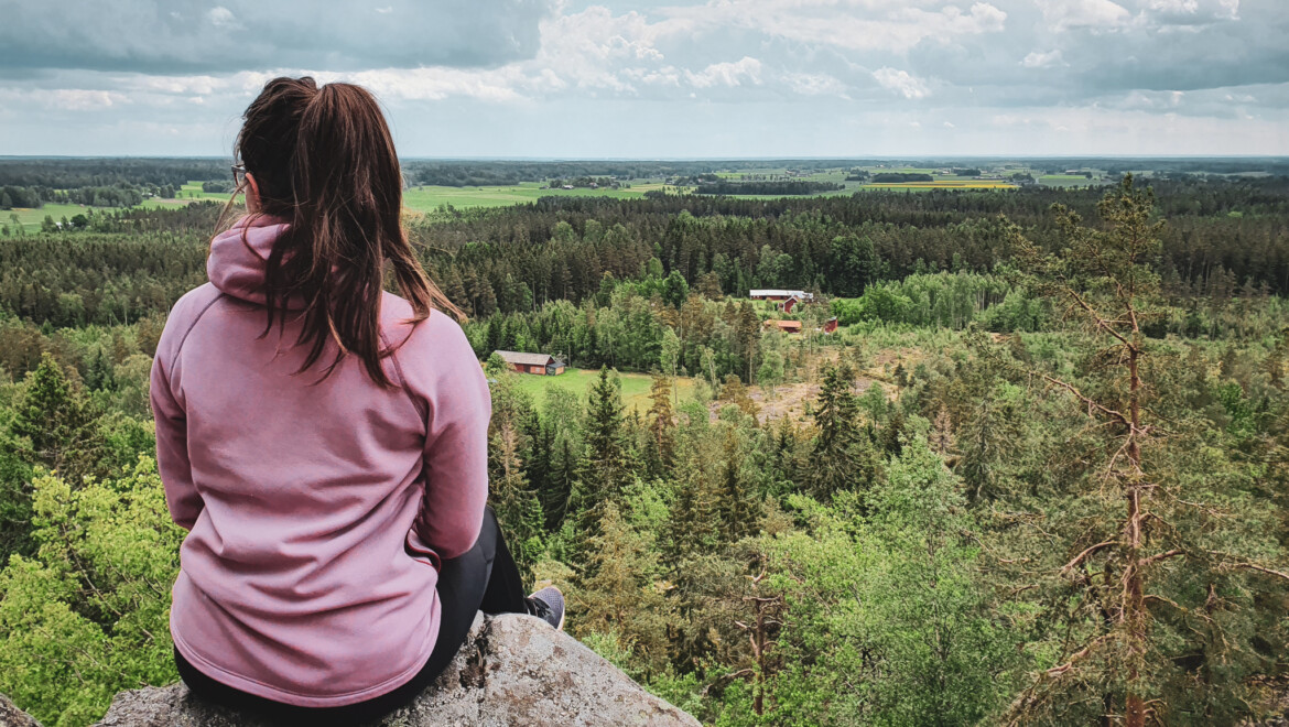 En kvinna med brunt hår i hästsvans och rosa huvtröja sitter på en klippa och tittar ut över utsikten med skog och ängar.