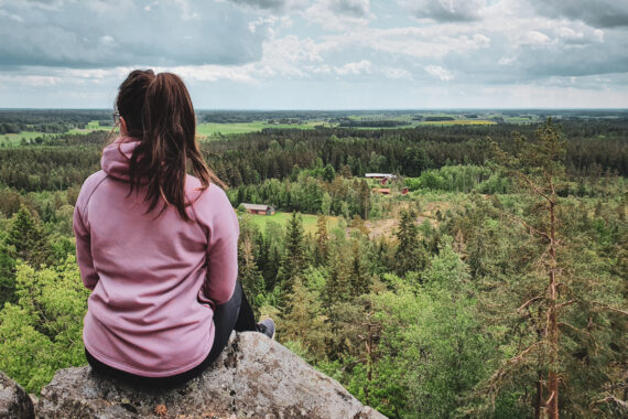 En kvinna med brunt hår i hästsvans och rosa huvtröja sitter på en klippa och tittar ut över utsikten med skog och ängar.