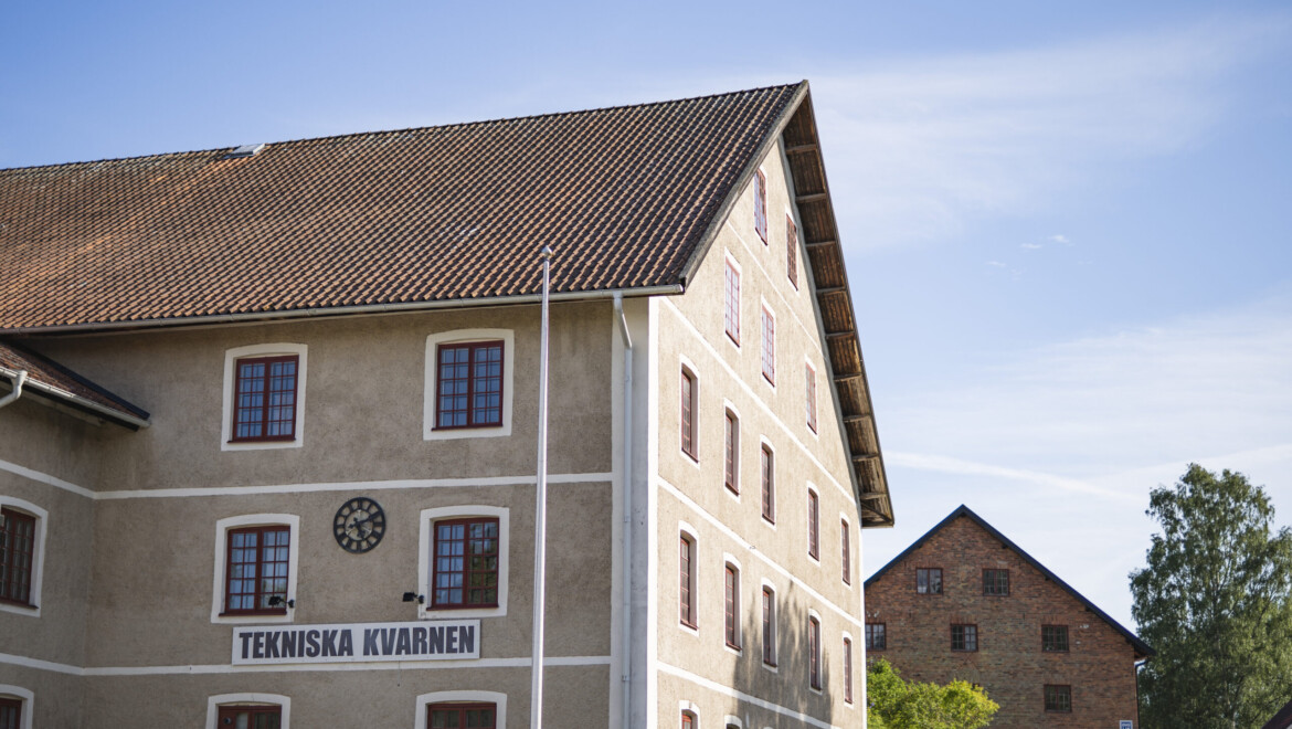 Övre hörnet av ett putsat hus i äldre uttryck med tegelhus och träd bakom.