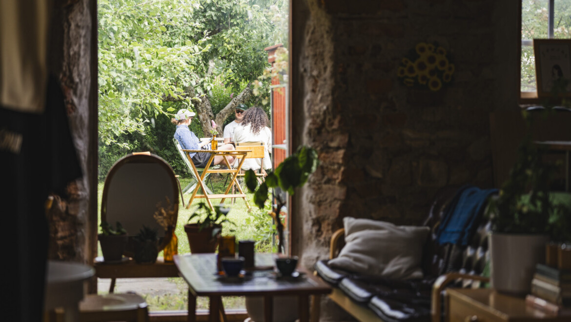 Tre personer syns på trädgårdsmöbler i grönska, genom ett fönster från ett rum med rustik inredning.