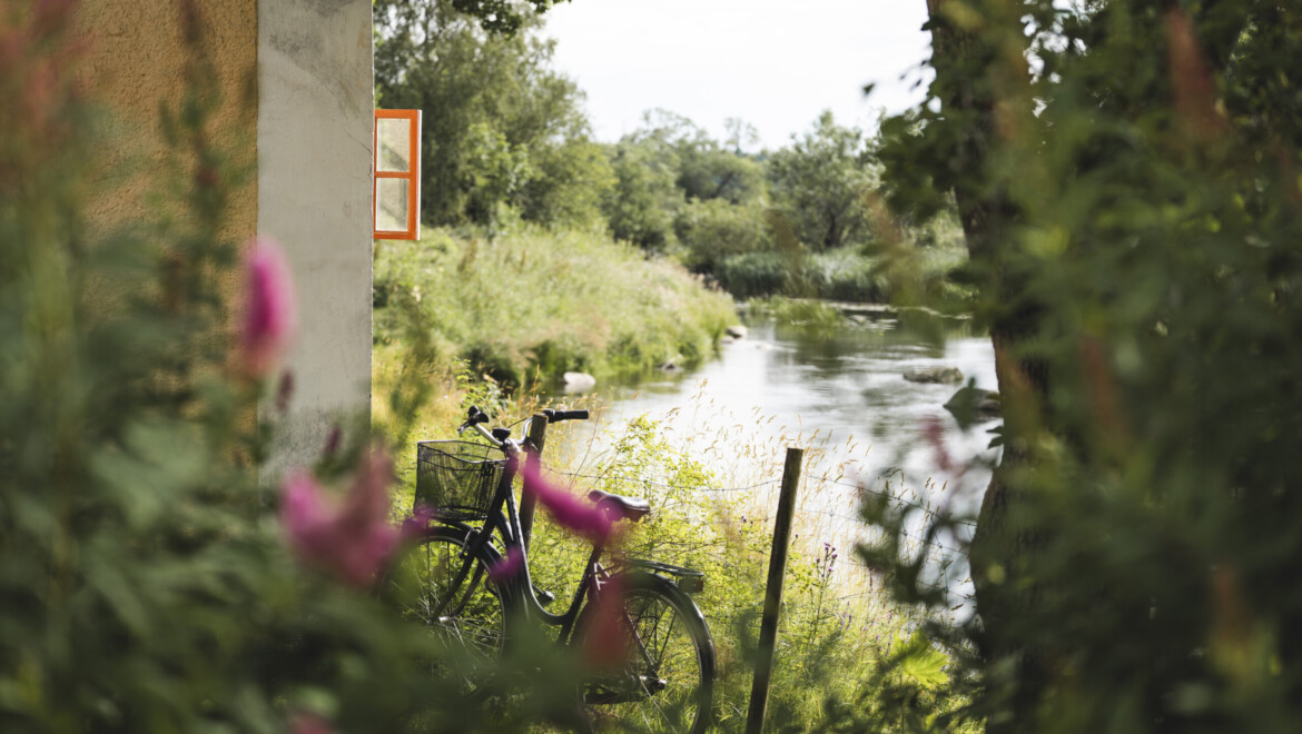 Cykel, gammalt hus och grönska står i förgrund till vattendrag.