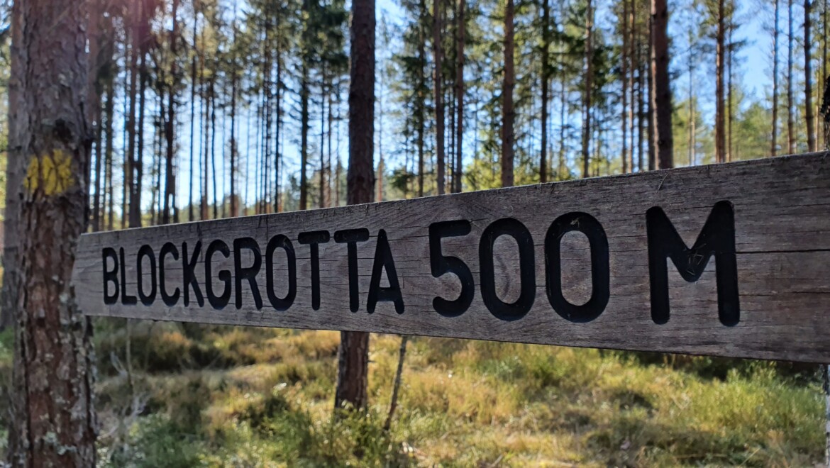 En skylt i trä med texten BLOCKGROTTA 500 M i svarta bokstäver. Runtom är det skog och mossa.