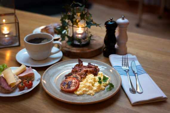 Äggröra, bacon och en grillad tomat ligger på en grå tallrik. Till vänster står en vit kaffekopp samt en assiett med en smörgås. Till höger ligger en kniv och en gaffel på en vit tygservett med blåa linjer.