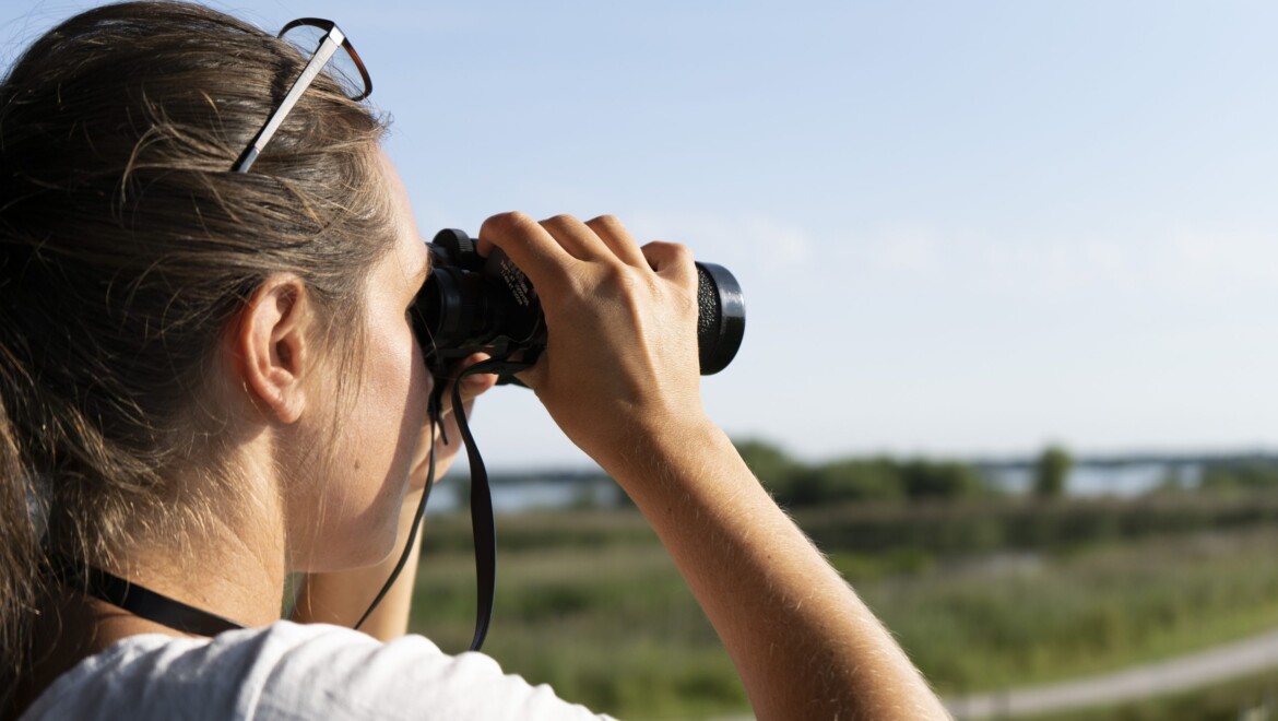 En kvinna i vit tröja håller en svart kikare framför ögonen. På huvudet sitter ett par glasögon fästa i håret.