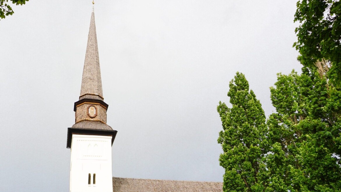 En vit, medeltida kyrka med spetsigt torn och träd i omgivningarna.
