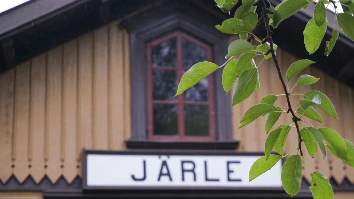 Ett gammalt stationshus i beige och brunt och röda fönsterspröjs. Huset har en skylt med texten JÄRLE på. En grön kvist hänger i förgrunden.