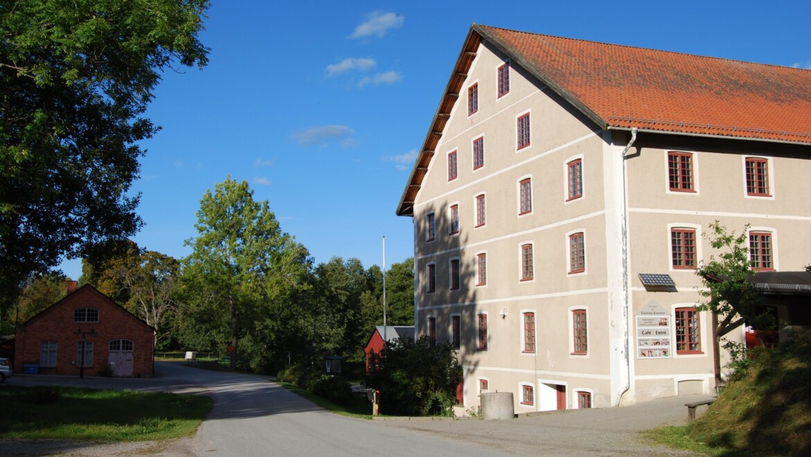 Ett sandfärgat hus med röda detaljer på fönstren. Huset ligger intill en väg med träd och buskar runtomkring.