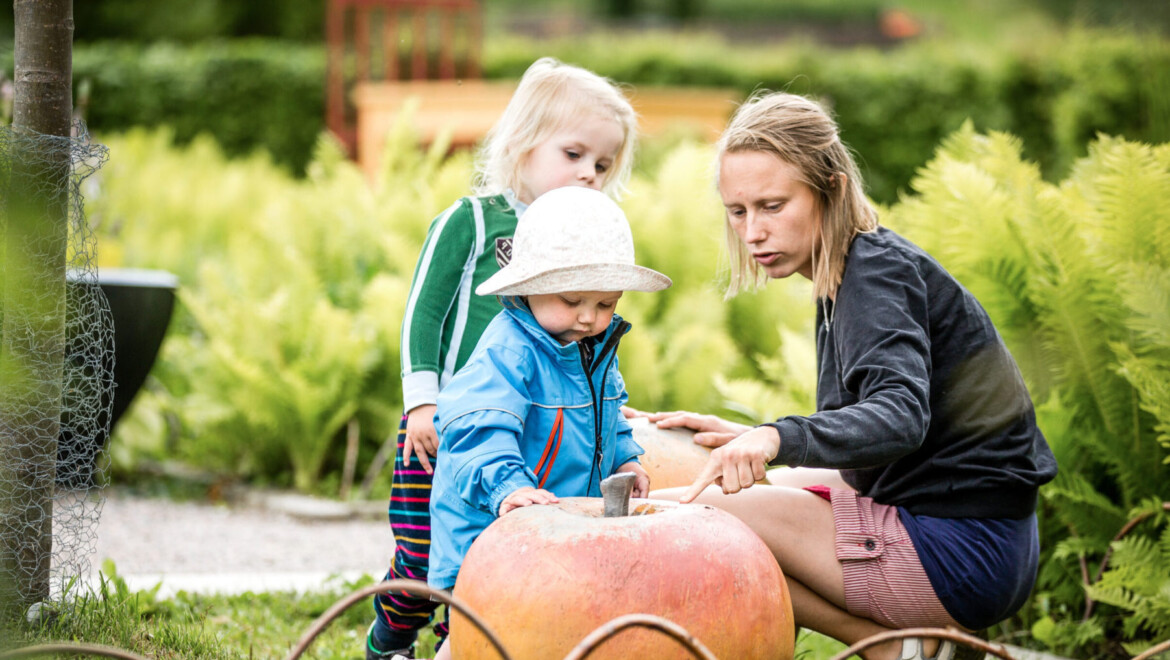 En kvinna med två små barn i en trädgård. Hon har handen på ett stort leksaksäpple.