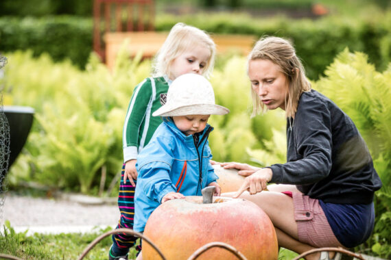En kvinna med två små barn i en trädgård. Hon har handen på ett stort leksaksäpple.