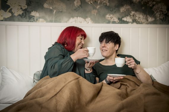 En kvinna i rött hår och en kvinna i svart hår har tekoppar i händerna och sitter tätt i en säng med brunt överkast.