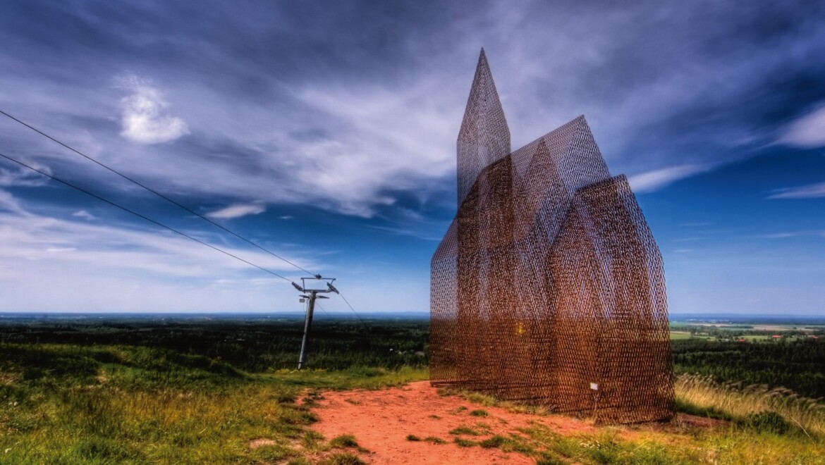 Ett konstverk som liknar en kyrka med torn, tillverkad av ett halvtransparent material. Står på en gräsbeklädd mark och himlen bakom är blå.