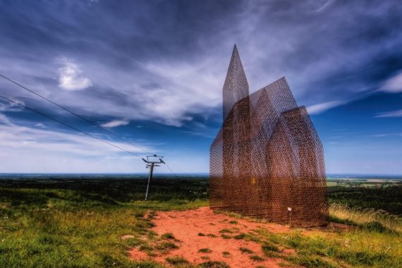 Ett konstverk som liknar en kyrka med torn, tillverkad av ett halvtransparent material. Står på en gräsbeklädd mark och himlen bakom är blå.