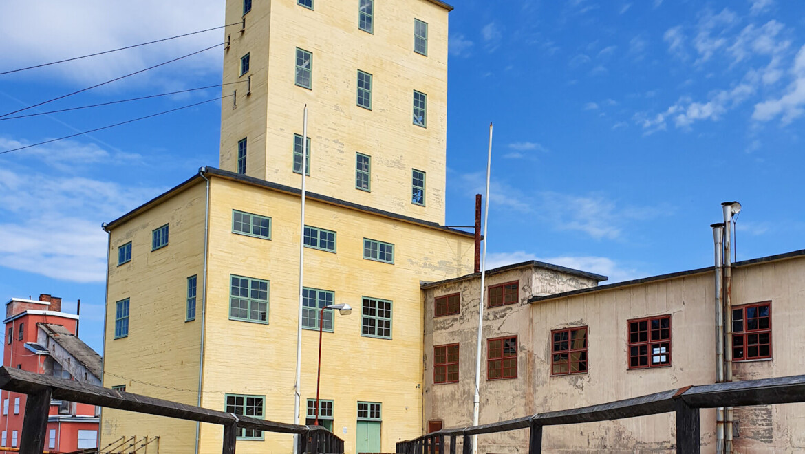 En gul industribyggnad med en järnvägsräls framför. Himlen är blå.