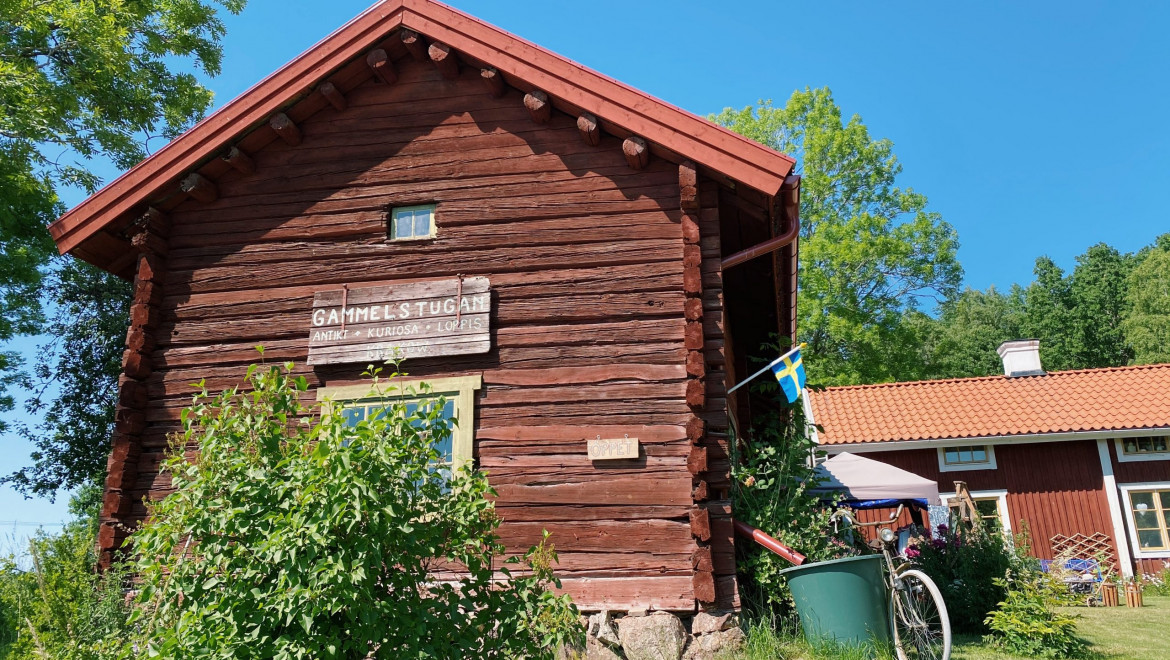 En röd timmerstuga med blå himmel och grön gräsmatta. Skylt med texten Gammelstugan hänger på stugans gavel.