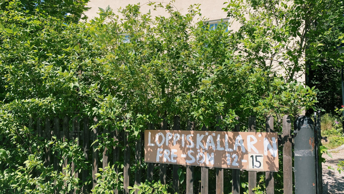 Ett ljusgult hus i bakgruden. Framför syns buskar och ett staket med skylten Loppiskällarn.