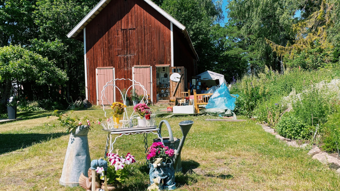 Ett rött skjul med grönt gräs och skog omkring. I förgrunden står stolar, vattenkannor och blommor utställda.