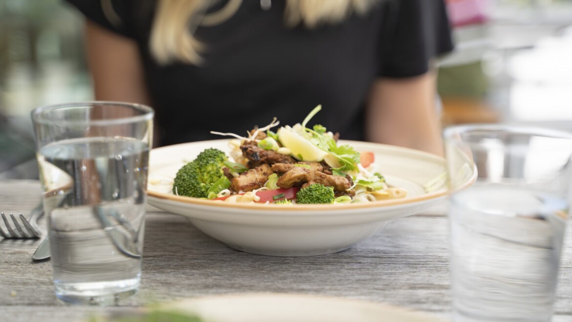 En vit, djup tallrik med kyckling och sallad. Två vattenglas och bestick. I bakgrunden syns en svart tröja och blonda hårslingor.