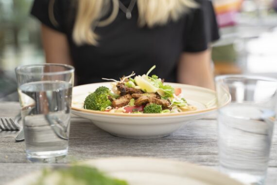 En vit, djup tallrik med kyckling och sallad. Två vattenglas och bestick. I bakgrunden syns en svart tröja och blonda hårslingor.