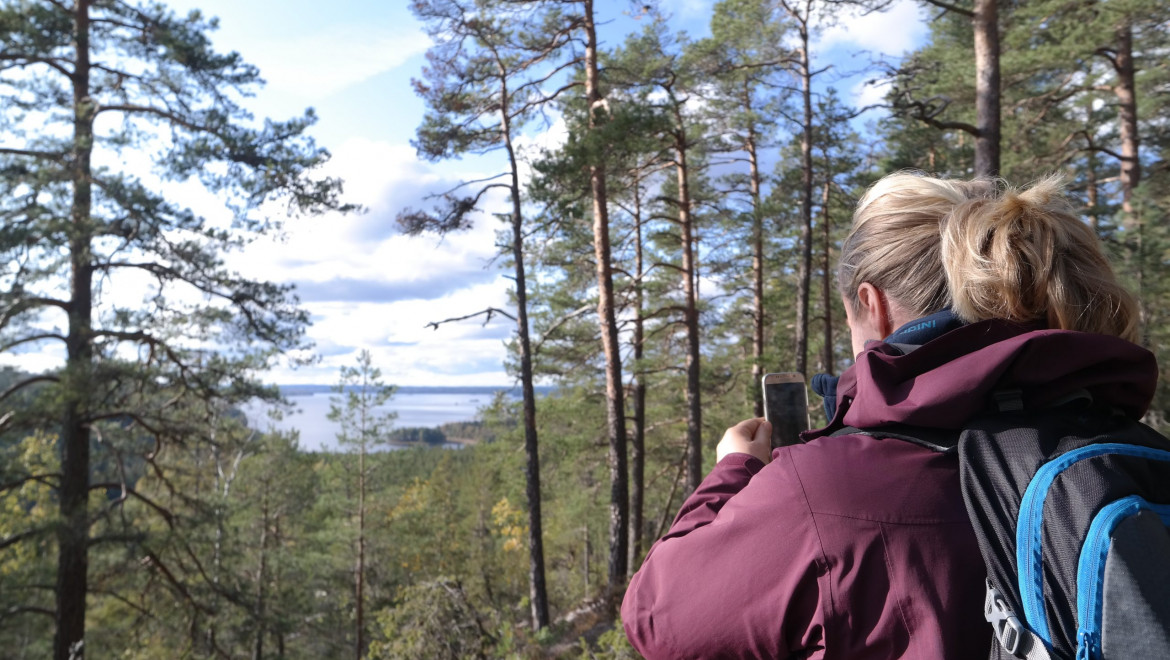 Utsikten mellan tallarna skymtas en sjö. En tjej med blond hästsvans och lila jacka fotar utsikten.