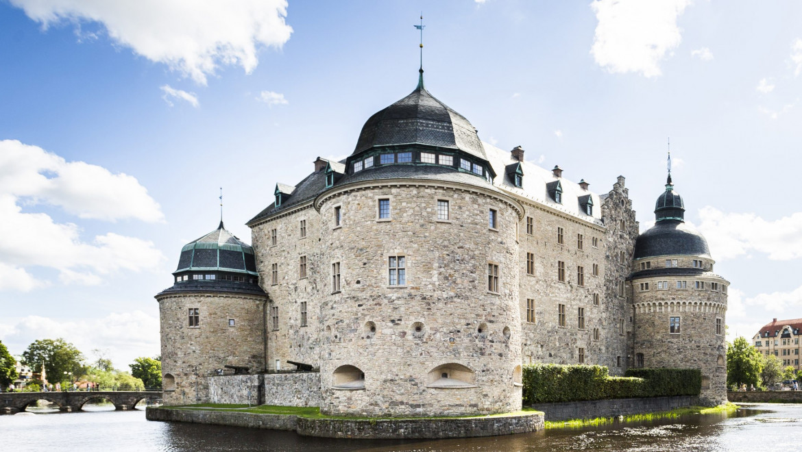 Ett sandfärgat slott med tre synliga torn och svart tak. Slottet står på en ö, omgiven av en å. Himlen är blå med några moln.