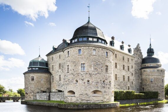 Ett sandfärgat slott med tre synliga torn och svart tak. Slottet står på en ö, omgiven av en å. Himlen är blå med några moln.