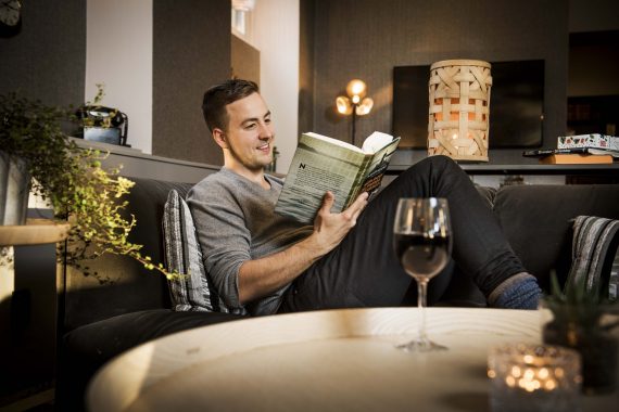 En person halvligger i en soffa och läser en bok. På bordet framför står ett vinglas.