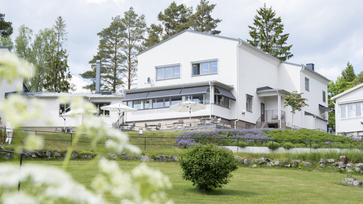 Ett vitt, stort hus i modern stil med gröna gräsmattor runtom, samt lila blommor i rabatter.
