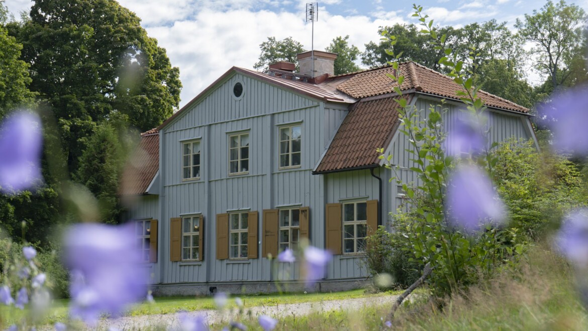 En grå trävilla med gula fönsterluckor är omgiven av gräs och blommor.