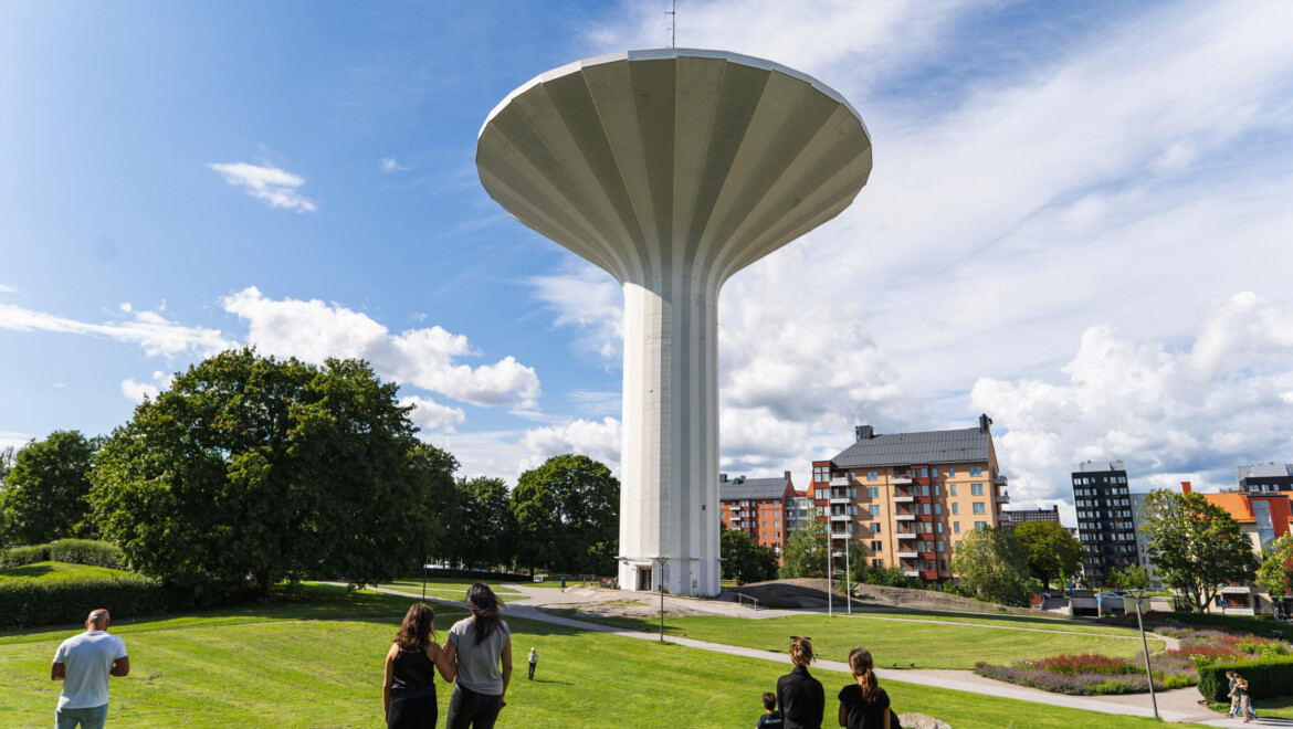 Stort vattentorn liknande en svamp, med grönt gräs och personer som går framför.