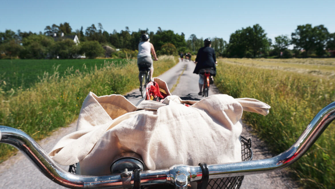 Ett cykelstyre med en vit kasse i korgen. Framför cykel syns en stig där två damer cyklar.