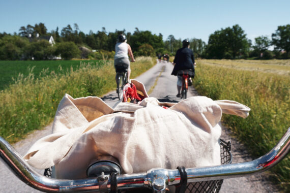 Ett cykelstyre med en vit kasse i korgen. Framför cykel syns en stig där två damer cyklar.