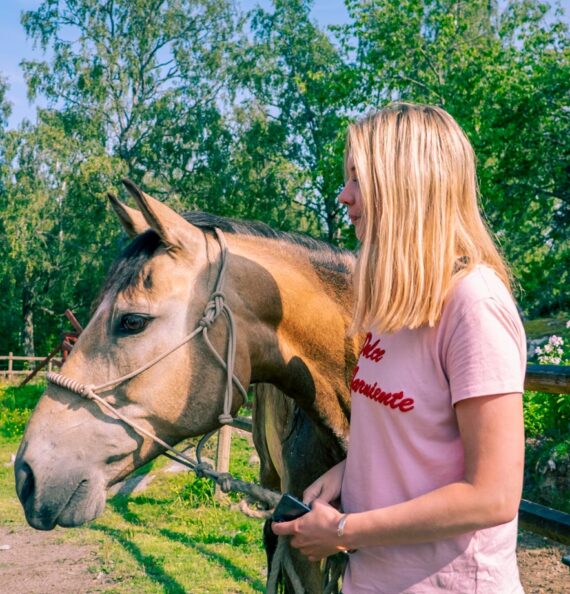 En kvinna i rosa tröja håller i tyglarna på en beigefärgad häst.