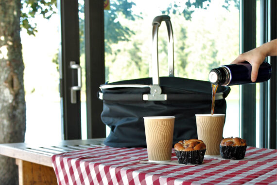 En rödrutig duk och svart picknickväska med två muggar och två bullar.
