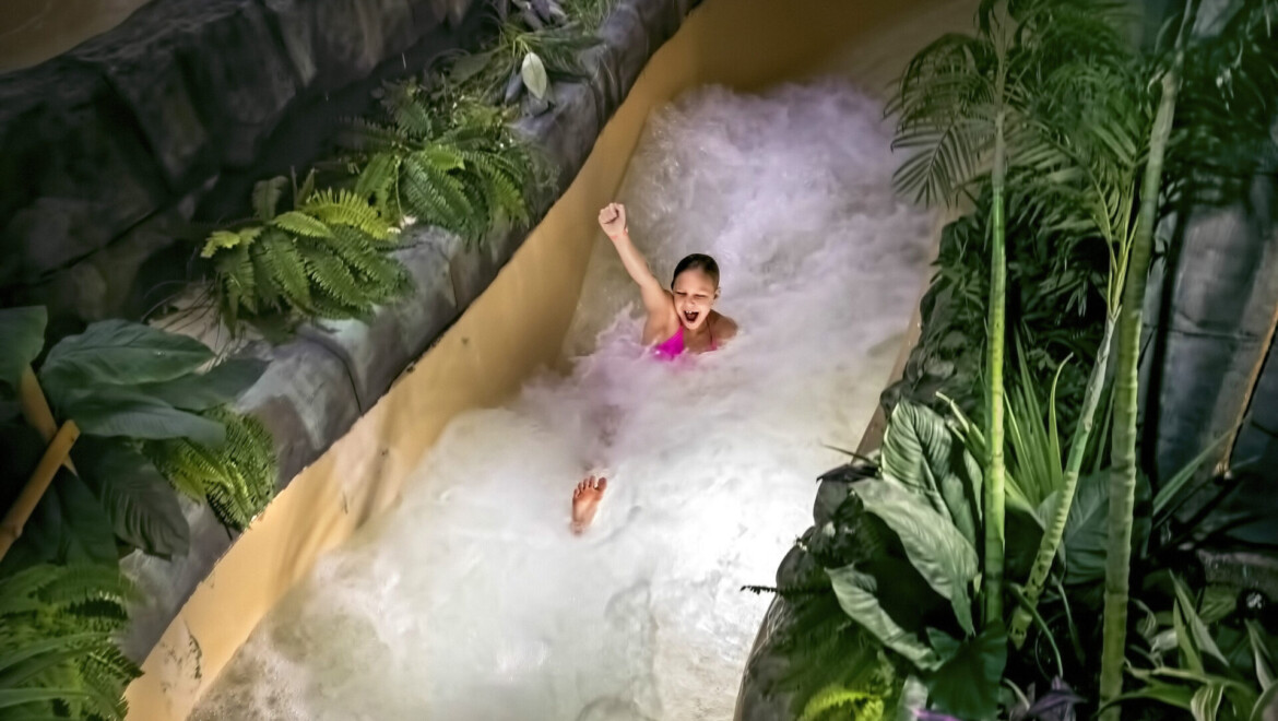 Glad tjej som åker en typ av vattenrutschbana omgiven av djungelliknande växtlighet.