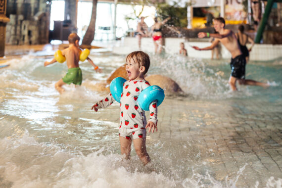 Ett barn med prickiga badkläder och blå armpuffar står i en barnpool och skrattar.