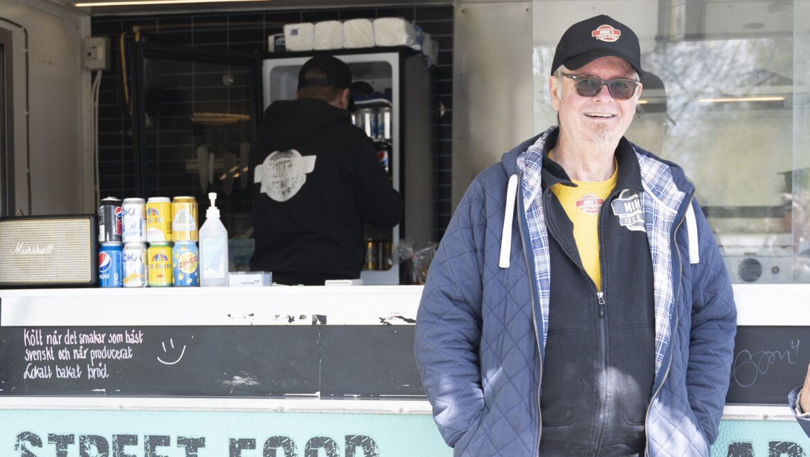 En man med keps och glasögon ser glad ut och står framför en foodtruck.