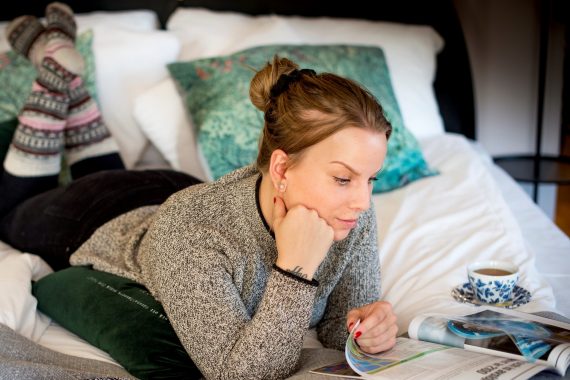 En person ligger på mage i en säng med raggsockor på sig och läser ett magasin.