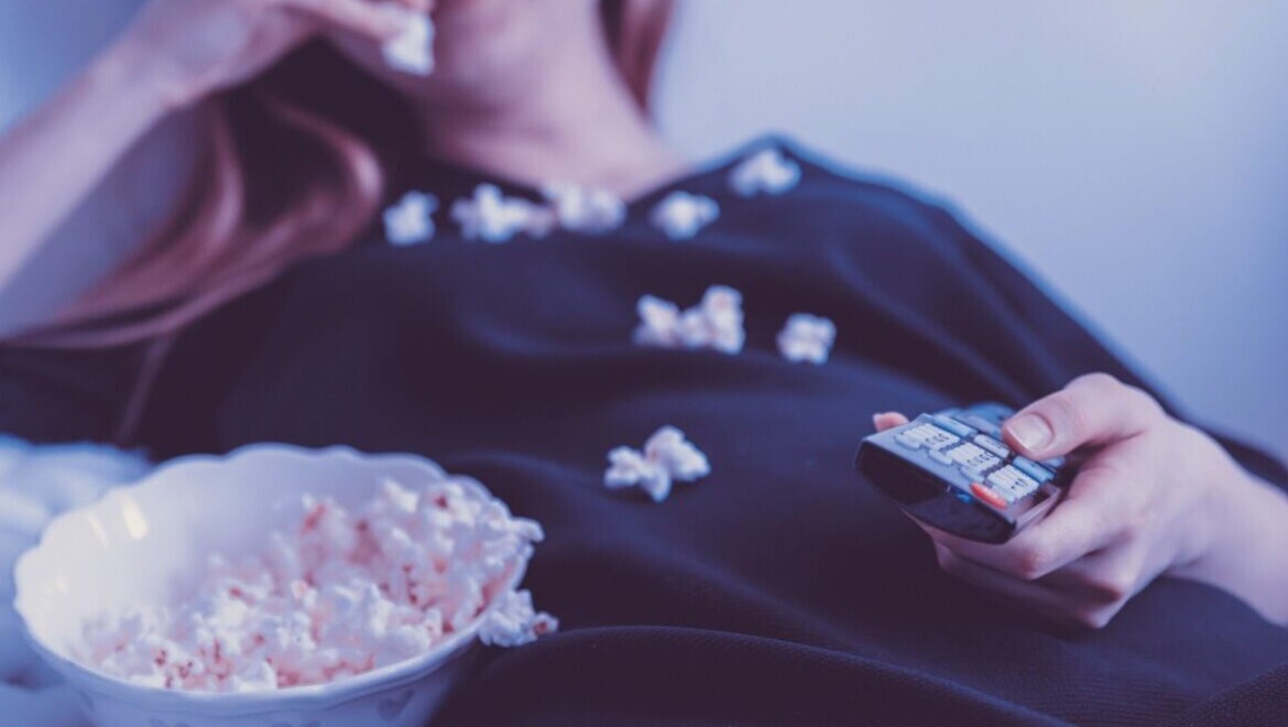 En kvinna halvligger och äter popcorn och har en TV-kontroll i handen.