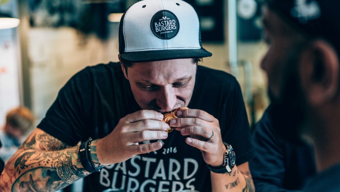 En man med tatueringar och svartvit keps äter en hamburgare med båda händerna.