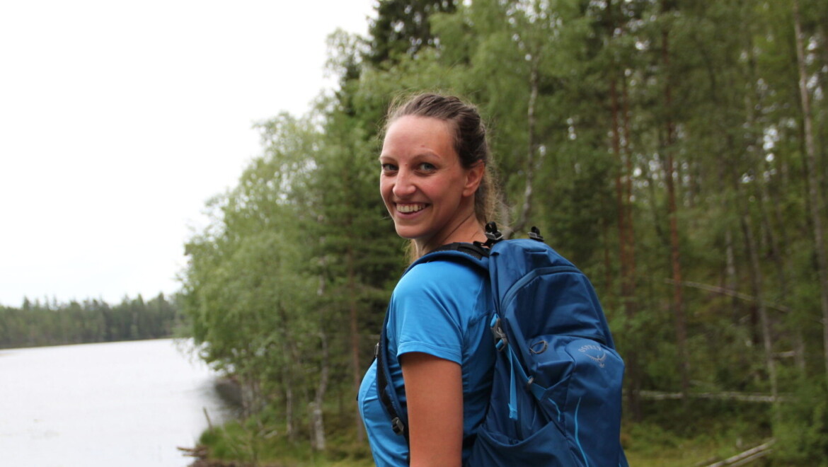 En kvinna med blå T-shirt och blå ryggsäck står i ett grönt landskap.