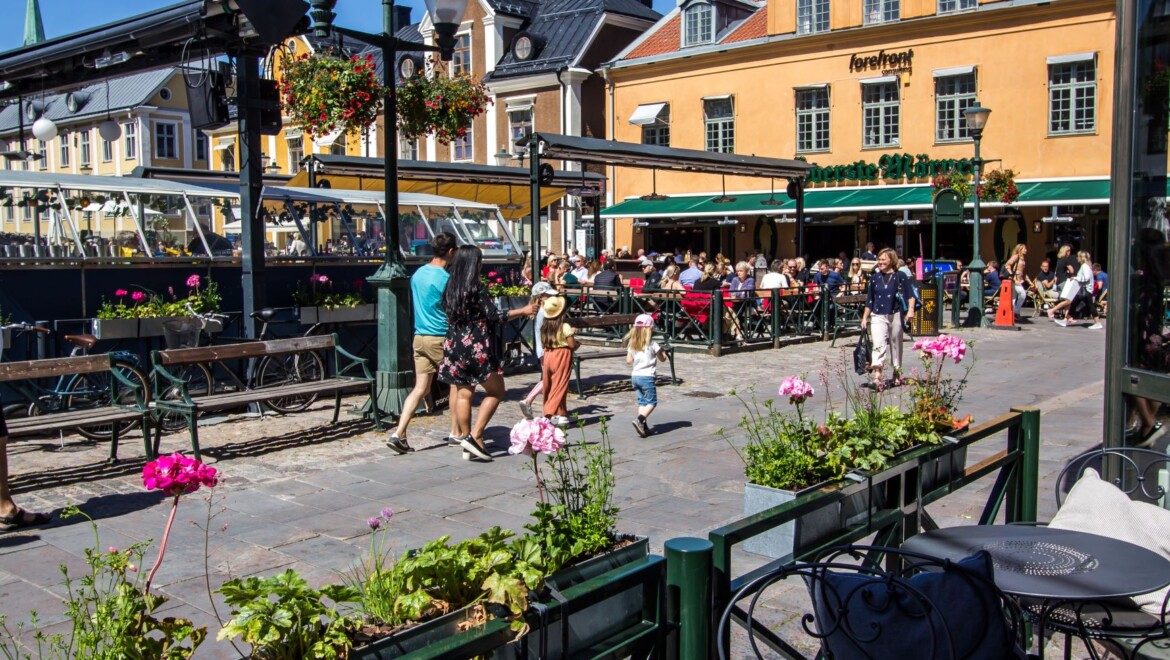 En uteservering med blomstrande blomlådor. Personer går på torget framför.