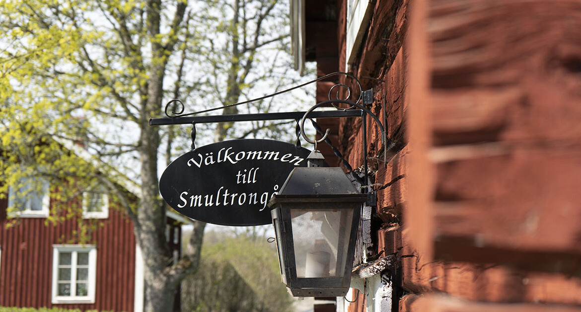 En svart skylt i gammeldags stil med texten Smultrongården sitter på en röd timmerbyggnad.