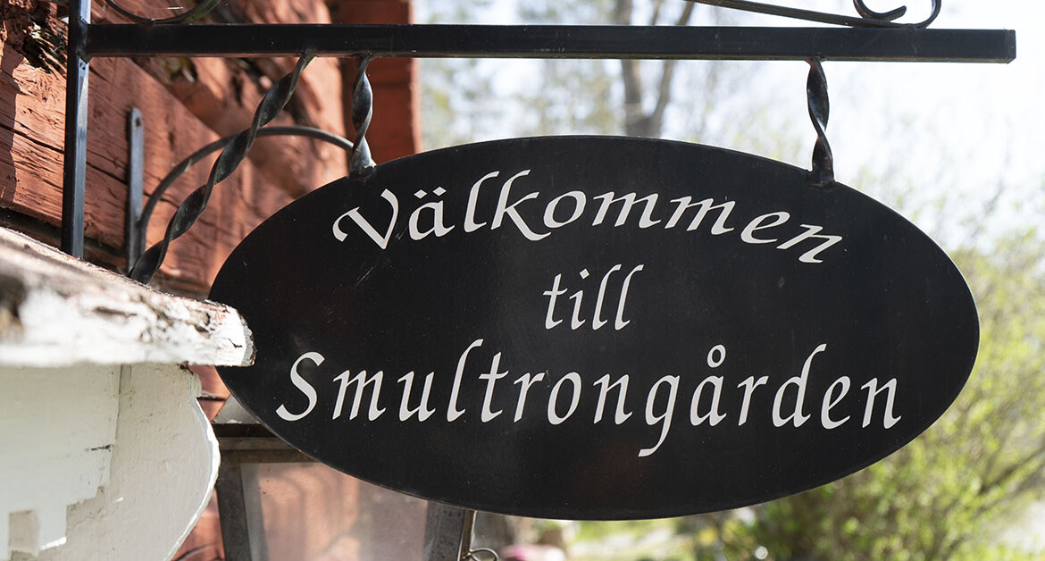 En svart skylt i gammeldags stil med texten Smultrongården sitter på en röd timmerbyggnad.