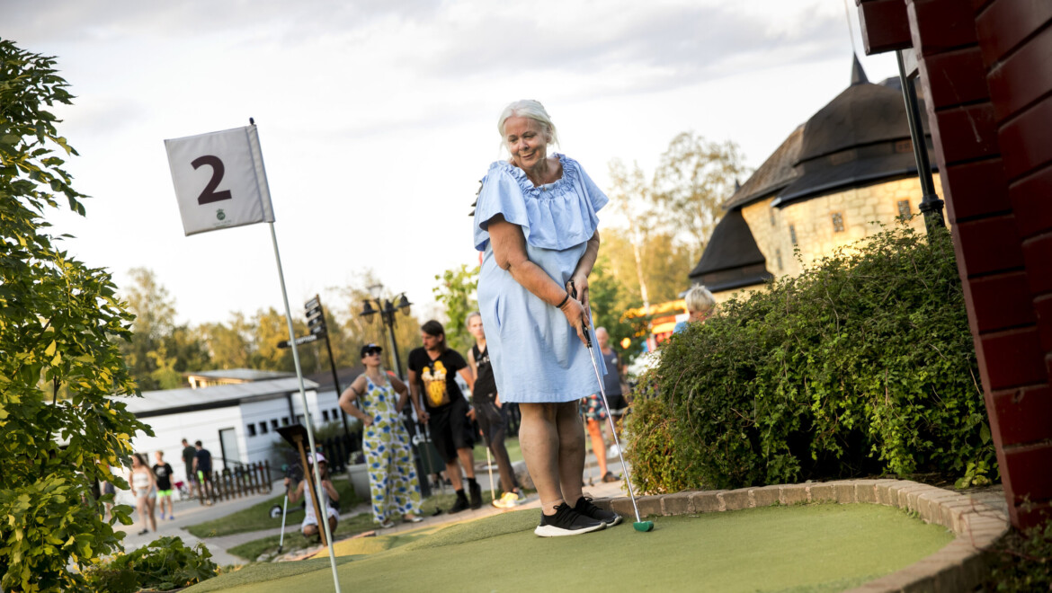 En äldre kvinna i ljusblå klänning spelar minigolf och ska precis putta bollen mot hålet.
