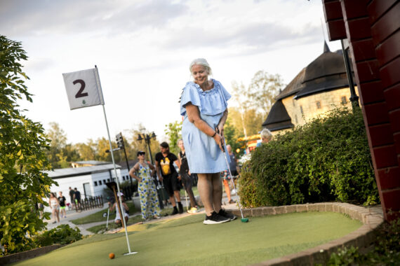 En äldre kvinna i ljusblå klänning spelar minigolf och ska precis putta bollen mot hålet.