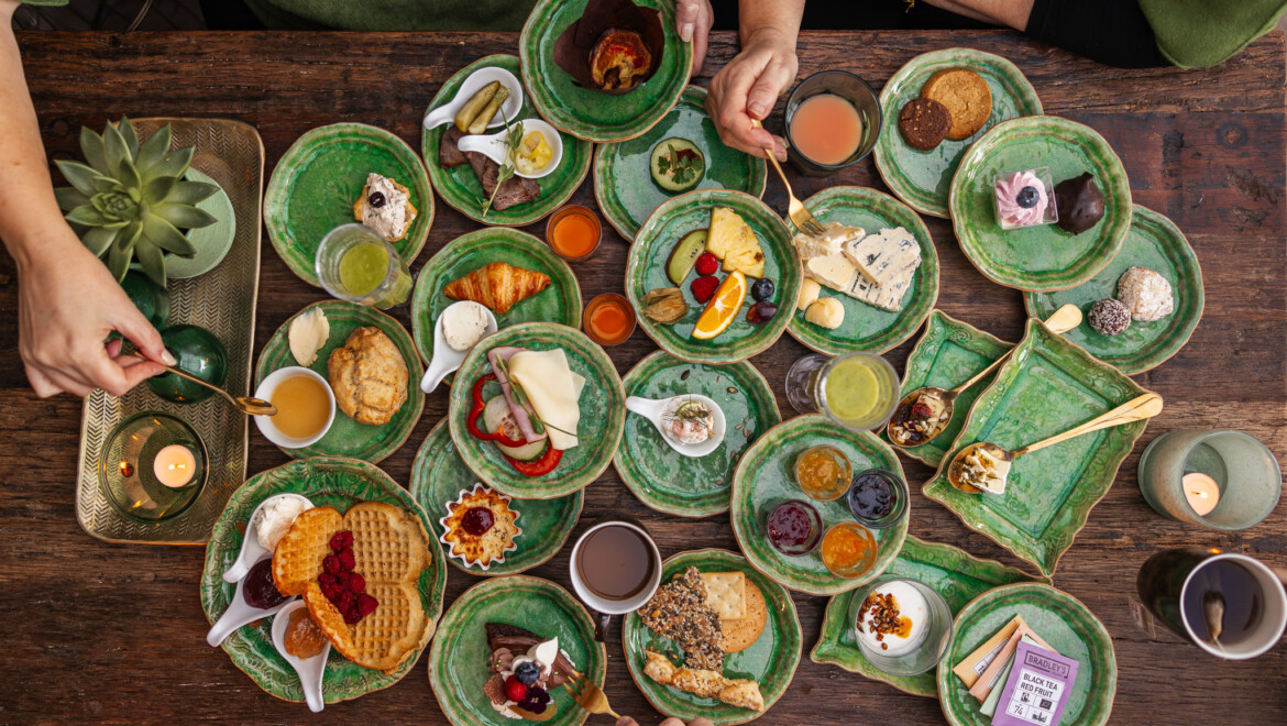 Många gröna tallrikar med frukosträtter på, sett ovanifrån. Händer som plockar bland porslinet.
