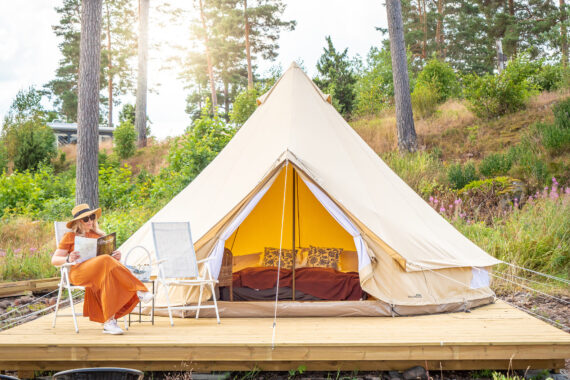 En kvinna med solhatt och solglasögon sitter i en stol på ett trädäck i en skog. Hon läser en tidning. På trädäcket står ett stort tält och i tältet syns en bäddad säng.