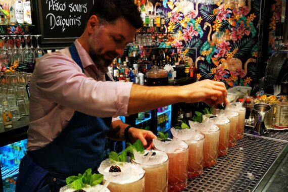En man förbereder många drinkar i en bar. Han står bakom baren och de ljusröda drinkarna står på baren. Han har på sig skjorta och förkläde. Bakom honom syns en färgglad tapet och en skylt med olika drinknamn på.