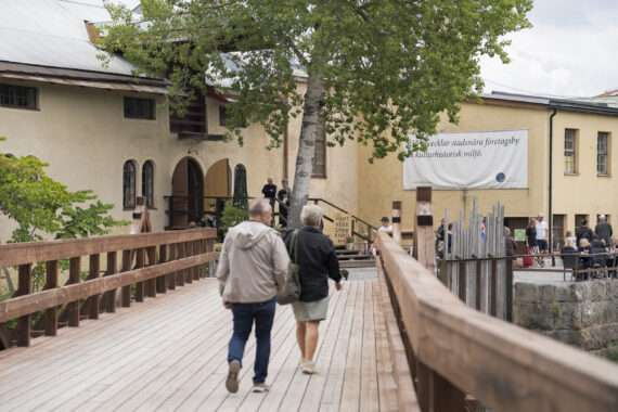 Två personer går på en träbro som leder fram till en stor gul byggnad. Vid byggnaden syns fler personer och på byggnaden sitter en stor, vit skylt med text på.