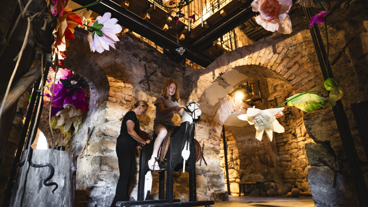 A girl is riding a rocking horse in a cave-like setting. A woman is standing next to her, smiling. Large paper flowers are attached to the walls.